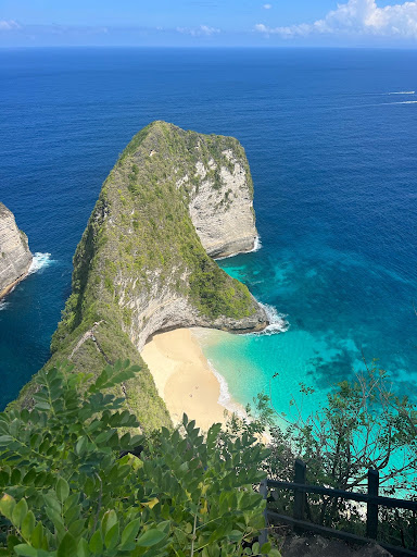 A dramatic green sea cliff rising above turquoise water and a sandy beach under a bright blue sky